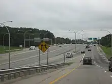 View of a three-lane collector/distributor road of a freeway; several green signs are visible in the distance. At the center of the image is a four-lane divided freeway that is separated from the distributor road by concrete barriers.