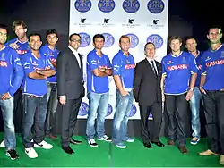 Nine players standing on a green carpet wearing a blue top and blue or black jeans. Along with them are two other men who are wearing black coats and pants.