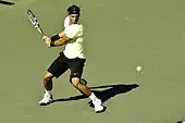 A brown-haired male tennis player with black shorts, a green shirt and a black headband swings a left-handed backhand on a hard court surface