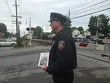 A police officer wearing a dark blue uniform faces left, holding a white pamphlet. Behind him a grade crossing gate is down, red lights are on and a train is approaching