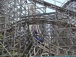 A blue train of the Rampage roller coaster is depicted reaching the bottom of the first drop to enter the slight left turn. From the viewers perspective, the train is heading to the right. The train is surrounded by winding wooden track and support structure, with several parts above the drop and in the background.