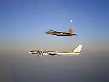 Aerial port view of two aircraft in flight, one on top of the other. The bottom aircraft is a four-engined propeller-driven aircraft, which is escorted by a jet fighter.
