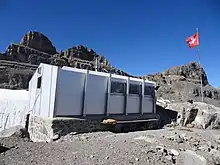 A white metal hut in the foreground, a mast with a Swiss flag to its right and rocky mountains in the background.