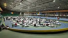 An indoor sports hall with people on camp beds inside a running track