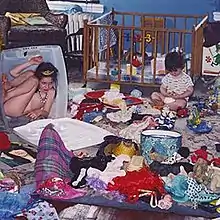 A photograph of two children in a room. On the left, a child in a tiara sits inside a clear plastic box; on the right, a child sits and plays in front of a wooden crib. The room is filled with various other children's items.