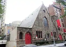 A brown stone triangular building with a square tower at the right on a city street. Its windows have pointed arches and there are similarly shaped red doors on either side. A large purple banner hanging from it has the word "Resurrection" on it