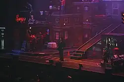 A man standing in front of the audience while in the middle of a concert, surrounded by the apartment-styled scenography and the rest of the musicians.