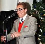 man making impression of George Burns between microphone and Christmas tree