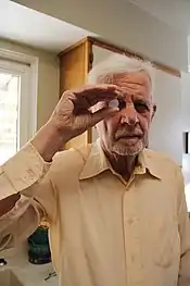 A man with white hair wearing a yellow collared shirt holding a small transparent in front of his right eye