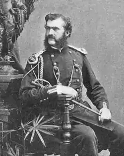 A monochrome photograph portrait of a man with light skin and dark hair, including a bushy muttonchop whiskers and moustache, the man seated, holding a thin sword at rest on his leg, wearing a military uniform decorated with epaulets, braid and buttons