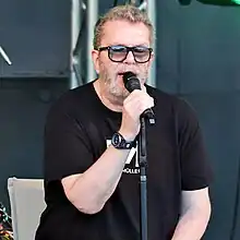 close-up of Richard Müller wearing a black print t-shirt and tinted sunglasses, sitting on a chair onstage, appearing to sing into a microphone