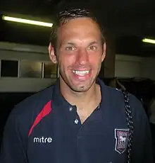 A man in a navy blue open-necked shirt with a crest and the word "mitre" on the chest, smiling.