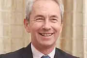 Official colour portrait photograph of Richard Stagg, smiling, standing in front of elaborate stonework