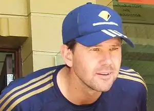 A man stands in front of a brick wall; he is wearing a dark blue cap with matching stripes, and a matching T-shirt. He is cleanshaven bar some stubble and has brown hair.