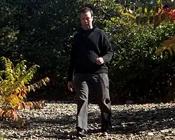 André Ristic wearing grey slacks and a black sweater, walking on a rocky trail, surrounded by trees and bushes