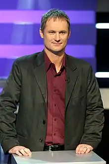 Medium-sized man with short brown hair wearing a maroon shirt and black jacket behind a desk on a purple background.