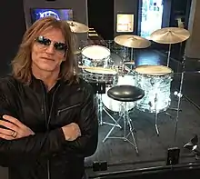 Rob Shanahan poses in sunglasses in front of a museum exhibit of Charlie Watts' drum kit