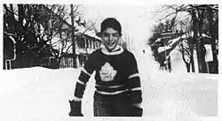 A young boy stands on a snow-covered street. He is wearing a dark-coloured sweater with a stylized maple leaf logo on the chest.
