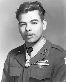 Head and shoulders of a bald man with dark beard, with a medal hanging from a blue ribbon around his neck.