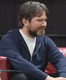 A man in his early 40s with brown hair and a brown beard looking to the right of the camera.