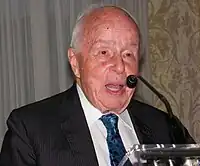 Rolf Noskwith, as an elderly man, speaking at a podium, with microphone partially obscuring his face.