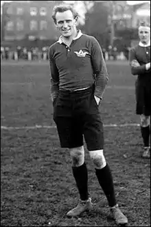Black and white photograph of Poulton in 1911. He is standing on a rugby field, wearing rugby sports clothing and smiling and looking into the camera.