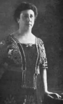 A white woman, standing, wearing a dark dress with a square neckline