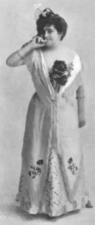 White woman standing in a light-colored gown with floral embellishments.