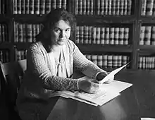 A young woman with fair skin and dark hair, seated at a table, holding papers, in front of a wall of bookshelves containing legal volumes