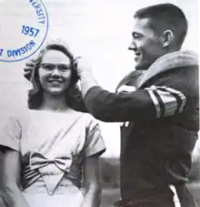 A young white woman and a young white man, both smiling; she is wearing glasses and a dress with a bow on the front; he is dressed for a football game; he is placing a tiara on her head