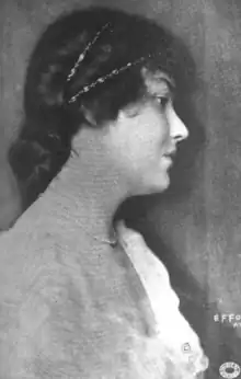 A young white woman in profile; her dark hair is styled in a low bun; she is wearing a white dress with a deep scoop necline