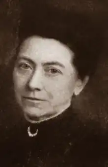 A middle-aged white woman with dark hair, wearing a dark dress with a high lace-trimmed collar