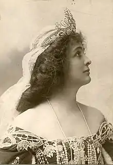 A white woman in profile, wearing a tiara and veil, with an off-the-shoulder gown trimmed in lace and pears, and a strand of pearls or beads.