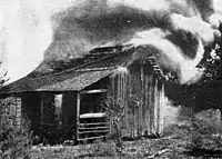 A black and white photograph of a crude wooden structure that could be a small shed, animal house, or hunting cabin with smoke pouring from it and flames visible in the door