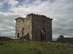 A tall stone built ruined square building. At the top are visible parts of the battlements. In the foreground a grassy slope with scattered stones leading up to the building. In the background white clouds with a small patch of blue sky.