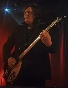 long-haired White male in dark suit, standing on musical stage holding a guitar, looking intently right of camera