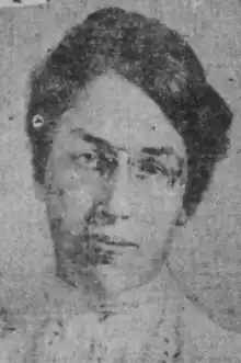 A young woman with light skin, dark hair parted and drawn back; she is wearing glasses and a high-collared white blouse.