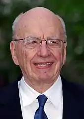 A head-and-shoulders photograph of an old, bald man with grey hair on the sides of his head. He is wearing spectacles and a white shirt with a blue tie and dark jacket.