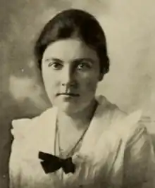 Young white woman with dark hair fastened back at the nape, wearing a white blouse with a frilled yoke and a dark bow at the chest