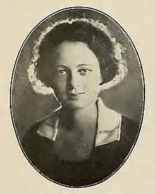 A young white woman with wavy dark hair, wearing a dark garment with a white collar; the lighting of the photograph lends a halo effect to her hair