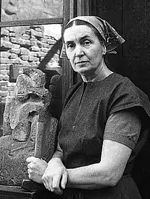 Black and white photo of Cravath posing next to a stone sculpture. She holds a mallet and is dressed in a black work dress. Her head is covered by a kerchief.
