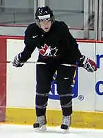 A Caucasian teenage ice hockey player. He stands crouched over towards the ice with his stick resting laterally on his thighs. He wears a black jersey with a stylized maple leaf logo.