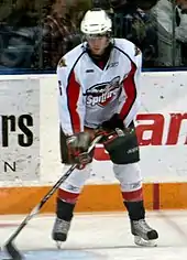 A teenage ice hockey player stickhandling a puck while standing still on the ice. His skates are shoulder-width apart and his eyes are downcast. He wears a white jersey with red and black trim, as well as a white, visored helmet.