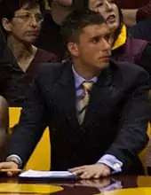 Waist high view of young man with short dark hair turning to one side wearing a blue shirt, dark blue suit and striped tie, two women visible behind him