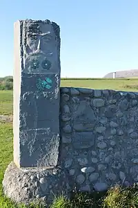 Section of cobblestone fence ending in a tall column with a bearded face carved into it. The column is decorated bird and fish figures as well as flowers.