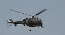 A small, camouflaged military helicopter, seen from the right and from below. South African markings can be seen on the aircraft's tail.