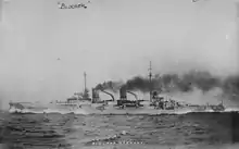 A light gray warship steaming at high speed; thick black smoke pours out of the two funnels.