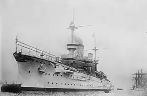 A large white warship bristling with guns sits in harbor; a tall sailing ship lies further aft in the background