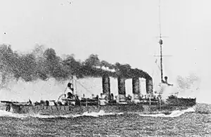 Waves crash against the bow of a cruiser as it steams ahead with large clouds of smoke coming out of all four of its funnels.