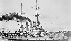 A large ship bristling with guns, thick black smoke pouring from its three funnels. Several other battleships are visible in the background.